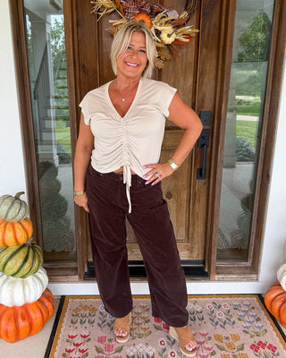A woman stands on a front porch wearing a cream ruched short-sleeve crop top and dark brown wide-leg pants, with stacked pumpkins on either side and a fall wreath above the door.