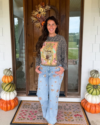 A woman stands on a front porch wearing a dark gray long-sleeve graphic sweatshirt with a colorful autumn-themed print and light blue distressed jeans with small embroidered motifs. The porch has a wooden door, white brick walls, fall wreath, and stacked pumpkins on both sides.