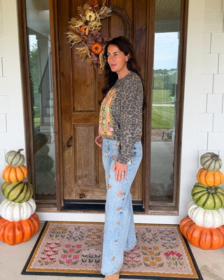 A woman stands on a front porch wearing a leopard-print cardigan over a colorful top and light blue jeans with floral embroidery. The porch has a wooden door with a fall wreath, stacked pumpkins on both sides, and a patterned doormat.
