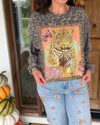 A woman stands on a front porch wearing a gray leopard-print long-sleeve shirt with a large colorful tiger graphic on the front, paired with light blue jeans decorated with small floral patches. The background shows a wooden door with a fall wreath, stacked pumpkins, and a patterned doormat.