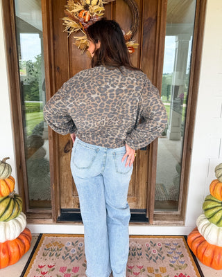 A woman stands with her back to the camera on a front porch, wearing a brown and beige leopard-print long-sleeve crop top and light-wash blue jeans. The porch has a wooden door with a fall wreath, a floral rug, and stacked pumpkins on both sides.