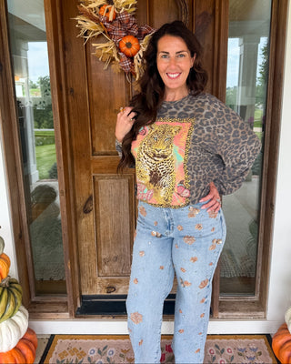 A woman stands on a front porch wearing a leopard-print long-sleeve top with a colorful graphic print on the front, paired with light blue jeans decorated with small floral patterns. She is posing in front of a wooden door with a fall wreath, with pumpkins visible on either side.