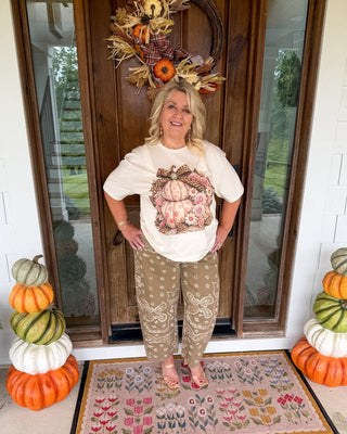 A woman stands on a front porch wearing a cream graphic t-shirt with a pumpkin print and beige patterned pants. She is positioned in front of a wooden door decorated with a fall wreath, with stacked pumpkins on both sides of the doorway.