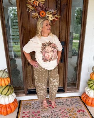 A woman stands on a front porch wearing a cream graphic t-shirt with a pumpkin design and tan patterned pants. She is positioned on a decorative doormat in front of a wooden door with autumn wreath decorations and stacked pumpkins on both sides.