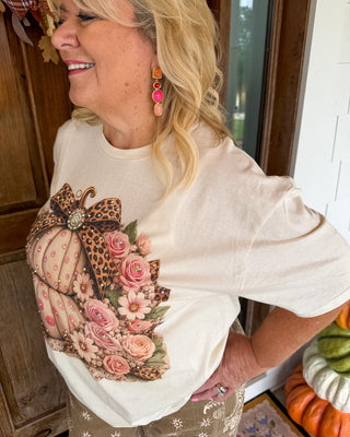 A woman wearing a cream-colored short-sleeve t-shirt with a large autumn graphic of a patterned pumpkin, pink roses, and a leopard print bow. She is standing in a home setting with fall decorations visible in the background.