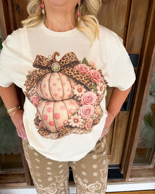 A woman wearing a cream-colored graphic t-shirt featuring a large autumn pumpkin design with pink roses and a leopard print bow, paired with tan patterned pants, standing in front of a wooden door.