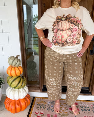 A woman stands on a front porch wearing a cream graphic T-shirt with a large floral pumpkin print and tan patterned pants. A stack of pumpkins sits to the left, and a patterned rug is visible beneath her feet.