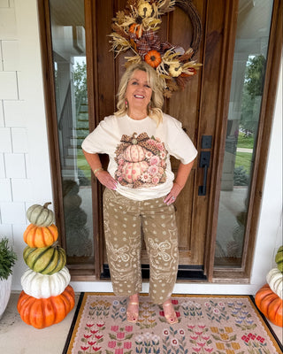 A woman stands on a front porch wearing a cream graphic T-shirt with a pumpkin and floral print, tan patterned pants, and pink heels. The porch has autumn decorations including stacked pumpkins, a wreath, and a patterned doormat.