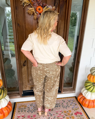 A woman stands with her back to the camera on a front porch, wearing a cream short-sleeve top and tan patterned pants. She is positioned in front of a wooden door with fall decorations, including a wreath and stacked pumpkins.