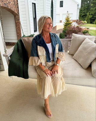 A woman sits on a porch swing wearing a cream and denim patchwork dress with a white top underneath. The porch has a concrete floor, a green knit blanket draped over the swing, and a white house in the background.