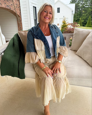 A woman sits on a beige outdoor sofa wearing a cream and blue patchwork ruffled jacket over a white top and a matching cream ruffled midi skirt. A green ribbed sweater is draped over the sofa beside her, with a house and trees in the background.
