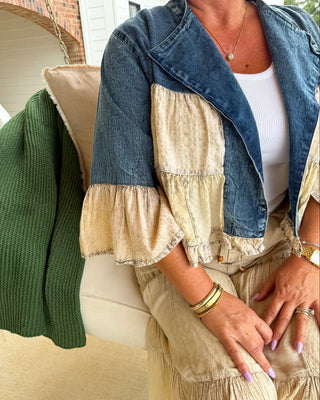 A person seated on a porch wearing a color-block denim jacket with cream ruffle sleeves and a matching cream tiered skirt. A green knit blanket is draped over a chair beside them.
