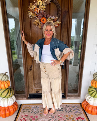 A woman stands on a front porch wearing a bohemian-style autumn outfit with a blue denim jacket, cream ruffled pants, and a white top. She is positioned in front of a wooden door decorated with a fall wreath, with stacked pumpkins on both sides and a floral doormat below.