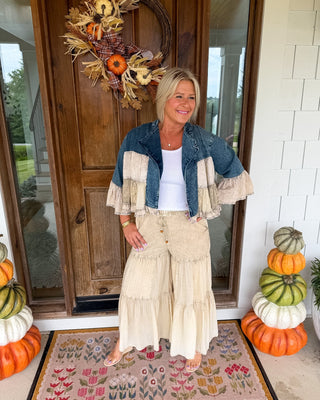 A woman stands on a front porch wearing a layered autumn outfit with a denim jacket featuring cream ruffle sleeves, a white top, and cream tiered wide-leg pants. She is positioned on a floral doormat in front of a wooden door decorated with a fall wreath, with stacked pumpkins on both sides.