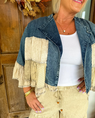 A woman wearing a patchwork denim jacket with beige ruffle panels, a white ribbed tank top, and beige fringed pants, standing in front of a rustic wooden door.