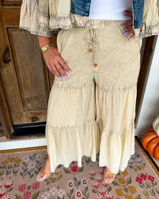 A person wearing wide-leg, tiered maxi pants in a light beige patchwork fabric, styled with a white top, a denim jacket, and clear slide sandals, standing on a floral rug.