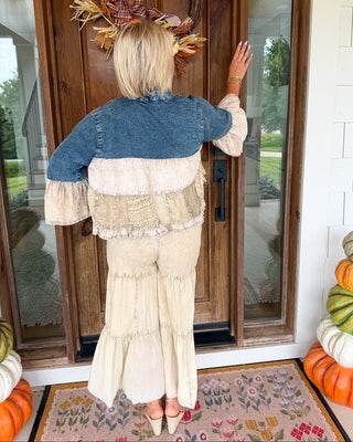 A woman stands with her back to the camera on a front porch, wearing a tiered cream maxi skirt and a denim-and-cream patchwork cropped jacket. She is touching a wooden door decorated with an autumn wreath, with stacked pumpkins on either side.