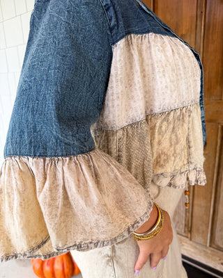 A close-up of a person wearing a bohemian-style patchwork blouse with a dark blue denim yoke and beige patterned ruffled sleeves, paired with a matching beige skirt. The photo is taken in a home setting with a wooden door and a small orange pumpkin visible in the background.