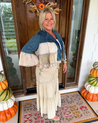 A woman stands on a front porch wearing a rustic patchwork outfit with a blue denim jacket and a long cream maxi skirt. The porch has autumn decorations including pumpkins, a floral doormat, and a wooden door.