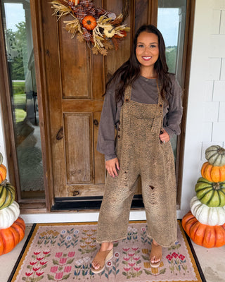 A woman stands on a front porch wearing a brown textured overall jumpsuit over a gray long-sleeve shirt. The porch has a wooden door with a fall wreath, stacked pumpkins on both sides, and a floral doormat.