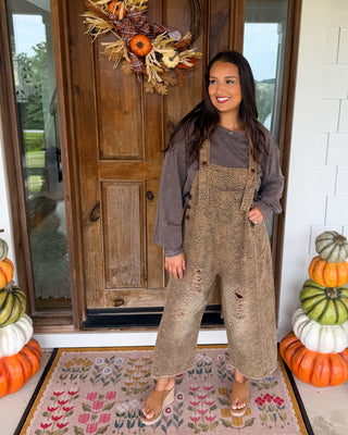 A woman stands on a front porch wearing a brown distressed overall jumpsuit over a dark gray long-sleeve shirt. The porch features a wooden door with a fall wreath, stacked pumpkins, and a floral doormat.
