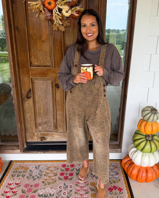A woman stands on a front porch wearing brown textured overall with a dark gray long-sleeve shirt, holding a mug. The porch has a wooden door with a fall wreath, a white brick wall, and a stack of pumpkins next to a potted plant.