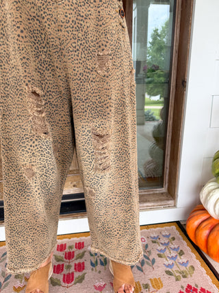 Close-up of a person wearing wide-leg beige pants with an allover leopard print and distressed rips on the legs, standing indoors near a glass door and patterned rug.