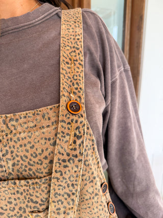 Close-up of a person wearing leopard-print denim overalls over a brown long-sleeve shirt, showing the patterned bib and shoulder strap with brown buttons.