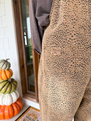 Close-up of a person wearing tan and brown leopard-print overalls with a gray long-sleeve top, standing near stacked pumpkins by a glass door.