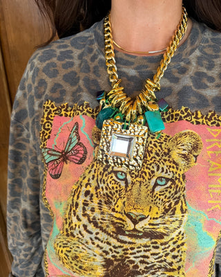 A close-up of a person wearing a chunky gold chain necklace with a large square pendant and turquoise accents, layered over a leopard-print sweatshirt with a colorful leopard graphic.