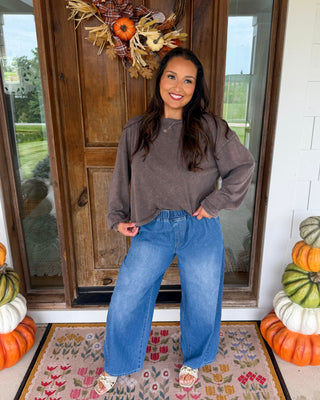 A woman stands on a front porch wearing a loose taupe-brown long-sleeve top and wide-leg blue jeans. The porch has a wooden door with a fall wreath, stacked pumpkins on both sides, and a floral rug underneath.