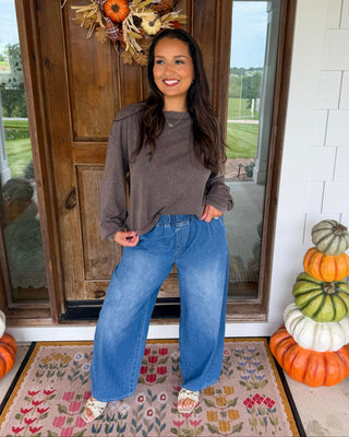 A woman stands on a decorated front porch wearing a brown long-sleeve top and wide-leg blue jeans. The porch has a wooden door with a fall wreath, a patterned rug, and stacked pumpkins on both sides.