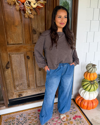A woman stands on a front porch wearing a brown long-sleeve cropped top and wide-leg blue jeans, with a floral doormat, wooden door, white brick wall, and stacked pumpkins nearby.