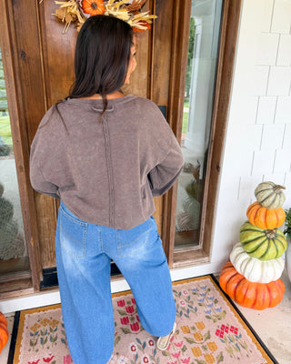 A woman viewed from behind wearing a brown cropped sweatshirt and blue jeans, standing on a decorated front porch with pumpkins and a wreath.