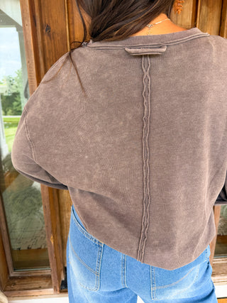 A person viewed from the back wearing a washed taupe-brown cropped sweatshirt and light blue jeans, standing in front of a wooden door.