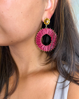 A close-up of a woman wearing a large circular drop earring. The earring features a vibrant magenta-pink pleated ring with gold-tone metal edging and a small round gold stud at the top.