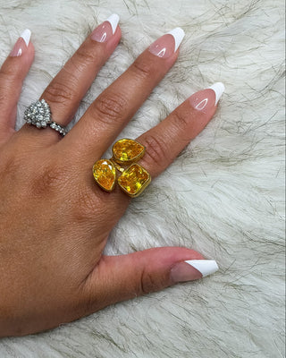A close-up of a hand with a French manicure wearing a large yellow gold ring with three marquise-cut yellow gemstones, alongside a silver ring on the opposite hand, set against a white faux fur background.