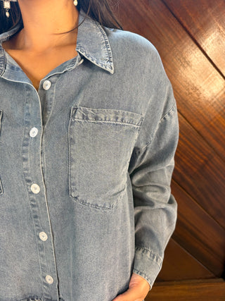 A close-up of a person wearing a light blue denim button-up shirt with a chest pocket, set against a dark wood-paneled background.