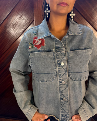 A woman wearing a light-wash denim jacket with a red and green floral embroidery on the left chest, standing against a dark wood paneled background.