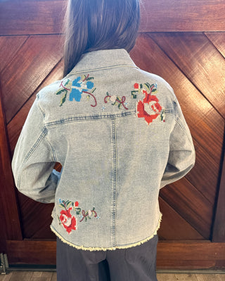 A person viewed from behind wearing a light-wash denim jacket with colorful floral embroidery on the back, paired with dark gray pants, standing in front of a wooden paneled wall.