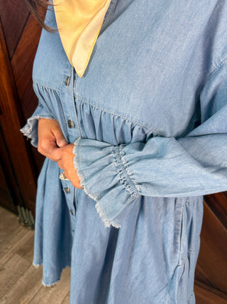 A close-up of a person wearing a light blue chambray button-front dress with gathered ruffle sleeves and a yellow collar lining, standing indoors near a wooden door.