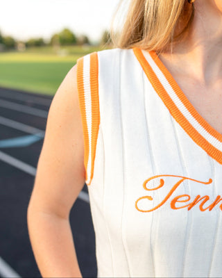 Close-up of a woman wearing a white sleeveless athletic top with orange and white striped trim and orange script lettering, photographed outdoors on a track field.