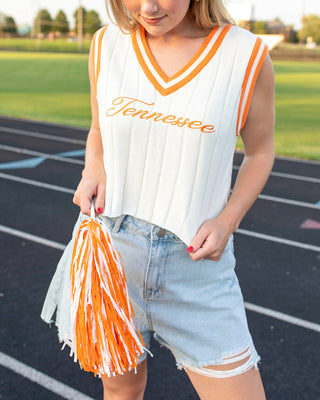 A woman standing on a track field wearing a white ribbed sleeveless V-neck top with orange trim and 'Tennessee' written across the chest, paired with light denim shorts and holding an orange and white pom-pom.