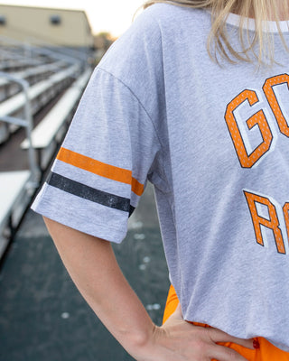 A close-up lifestyle shot of a person wearing a heather gray graphic t-shirt with orange and black sleeve stripes, paired with bright orange pants, standing on an outdoor bleacher deck.