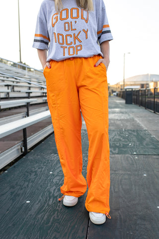 A person standing outdoors on a walkway wearing bright orange wide-leg pants, a light gray graphic T-shirt with orange lettering, and white sneakers.