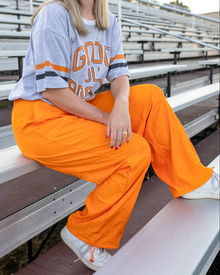 A woman sitting on outdoor stadium bleachers wearing a light blue and orange 'GOOD JOY' graphic t-shirt and bright orange wide-leg cargo pants.